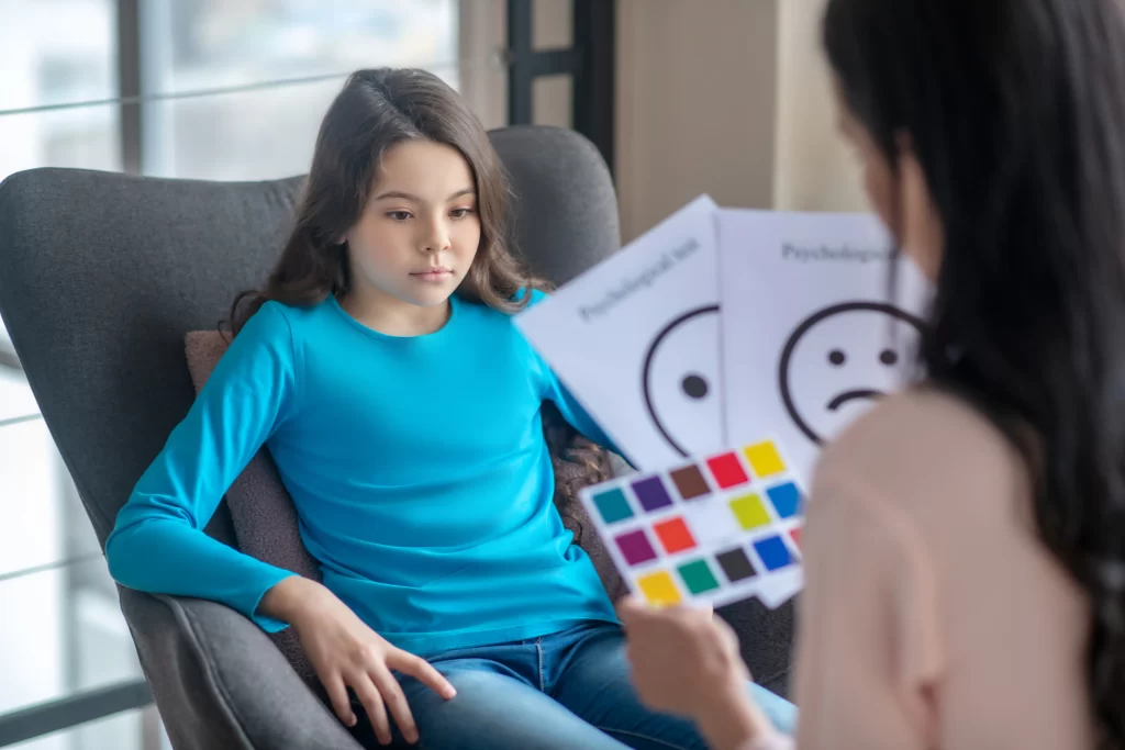 Una joven participa en una sesión de evaluación de psicologia infantil. Se observa a una profesional mostrando tarjetas con expresiones faciales y una paleta de colores.